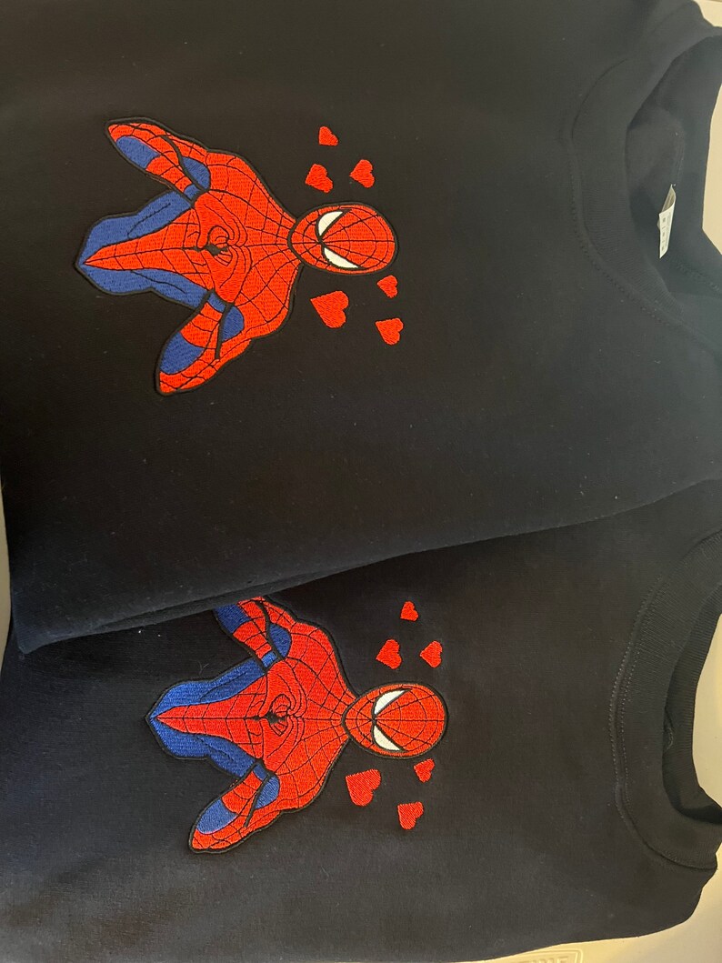 Puede incluir: Dos sudaderas negras con una imagen bordada de Spiderman con corazones a su alrededor, en rojo y azul.