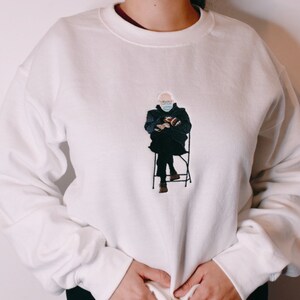 May include: White sweatshirt with an embroidered image of a person wearing a face mask and sitting on a chair.
