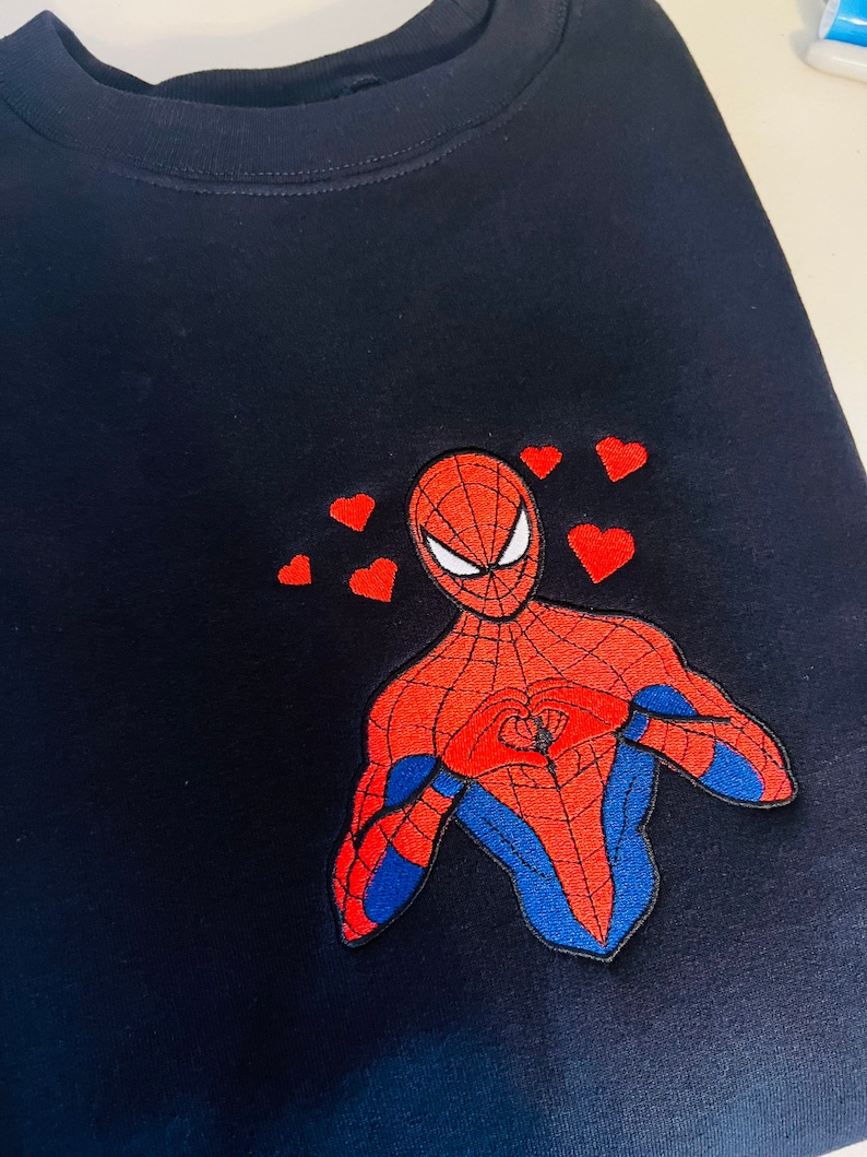 Puede incluir: Camiseta negra con una imagen bordada en rojo y azul de Spiderman haciendo un coraz&oacute;n con las manos. Hay varios corazones rojos peque&ntilde;os alrededor de Spiderman.