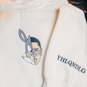 May include: White sweatshirt with an embroidered image of Bugs Bunny and a man's face. The sleeve has the text "YHLQMDLG" embroidered in black.