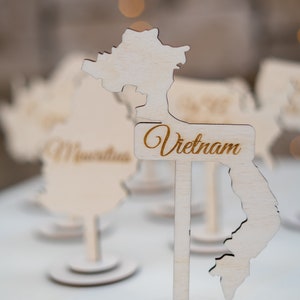 K&ouml;nnte beinhalten: Tischdekoration aus Holz in Form von Vietnam, mit dem Namen des Landes in Schreibschrift. Die Dekoration ist aus hellem Holz gefertigt und steht auf einem runden Sockel. Weitere &auml;hnliche Dekorationen sind im Hintergrund zu sehen.