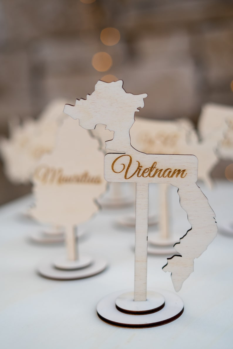 K&ouml;nnte beinhalten: Tischdekoration aus Holz in Form von Vietnam, mit dem Namen des Landes in Schreibschrift. Die Dekoration ist aus hellem Holz gefertigt und steht auf einem runden Sockel. Weitere &auml;hnliche Dekorationen sind im Hintergrund zu sehen.
