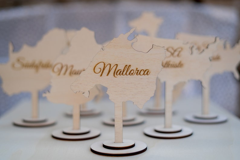 K&ouml;nnte beinhalten: Holz-Namensschildhalter in Form von Inseln, mit goldfarbenen eingravierten Namen. Einer tr&auml;gt die Aufschrift "Mallorca". Jeder hat einen runden Sockel und einen d&uuml;nnen Stiel.