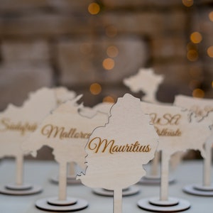 K&ouml;nnte beinhalten: Holz-Namenskartenhalter in Form von L&auml;ndern, mit dem Namen "Mauritius" in goldener Schrift. Die Halter stehen auf wei&szlig;en St&auml;ndern mit dunkelbraunen Basen, vor einem verschwommenen Hintergrund.