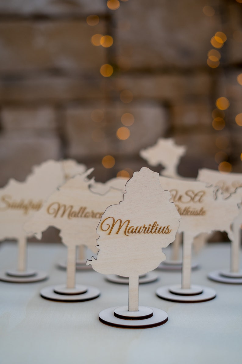 K&ouml;nnte beinhalten: Holz-Namenskartenhalter in Form von L&auml;ndern, mit dem Namen "Mauritius" in goldener Schrift. Die Halter stehen auf wei&szlig;en St&auml;ndern mit dunkelbraunen Basen, vor einem verschwommenen Hintergrund.