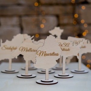K&ouml;nnte beinhalten: Holz-Namensschildhalter in Form von Kontinenten und L&auml;ndern, mit Namen wie "Mauritius" in eleganter Schrift graviert. Jeder Halter steht auf einem kleinen, runden Sockel. Der Hintergrund ist verschwommen mit warmen, goldenen Bokeh-Lichtern.