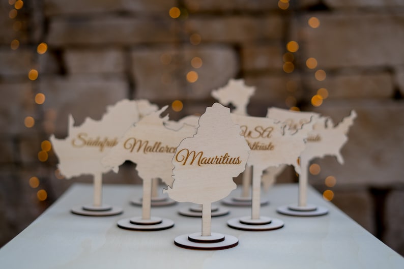 K&ouml;nnte beinhalten: Holz-Namensschildhalter in Form von Kontinenten und L&auml;ndern, mit Namen wie "Mauritius" in eleganter Schrift graviert. Jeder Halter steht auf einem kleinen, runden Sockel. Der Hintergrund ist verschwommen mit warmen, goldenen Bokeh-Lichtern.