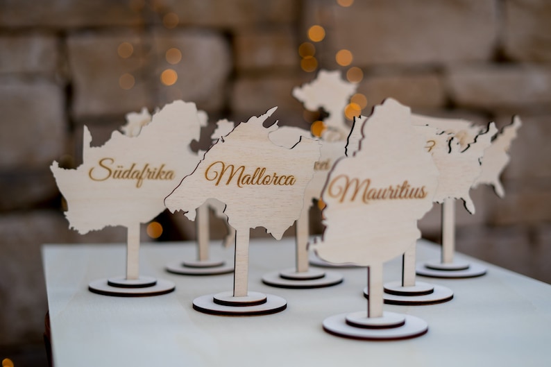 K&ouml;nnte beinhalten: Holz-Namensschilder in Form von L&auml;ndern, mit Namen wie "Mallorca" und "Mauritius" in goldener Schrift. Jede Karte steht auf einem kleinen, runden Sockel, ideal f&uuml;r Veranstaltungen.