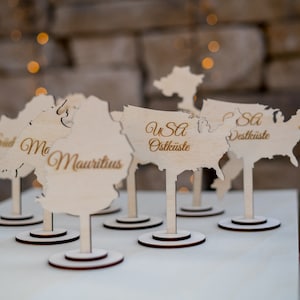 K&ouml;nnte beinhalten: Tischkarten aus Holz in Form von Kontinenten und L&auml;ndern, darunter die USA. Die Karten haben eine helle Holzoberfl&auml;che mit dunkelbraunen Sockeln und Stielen. Text in Gold identifiziert die Orte.