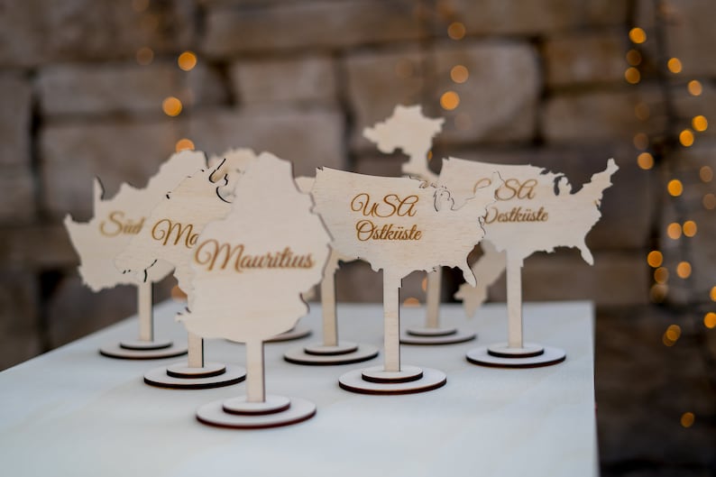 K&ouml;nnte beinhalten: Tischkarten aus Holz in Form von Kontinenten und L&auml;ndern, darunter die USA. Die Karten haben eine helle Holzoberfl&auml;che mit dunkelbraunen Sockeln und Stielen. Text in Gold identifiziert die Orte.