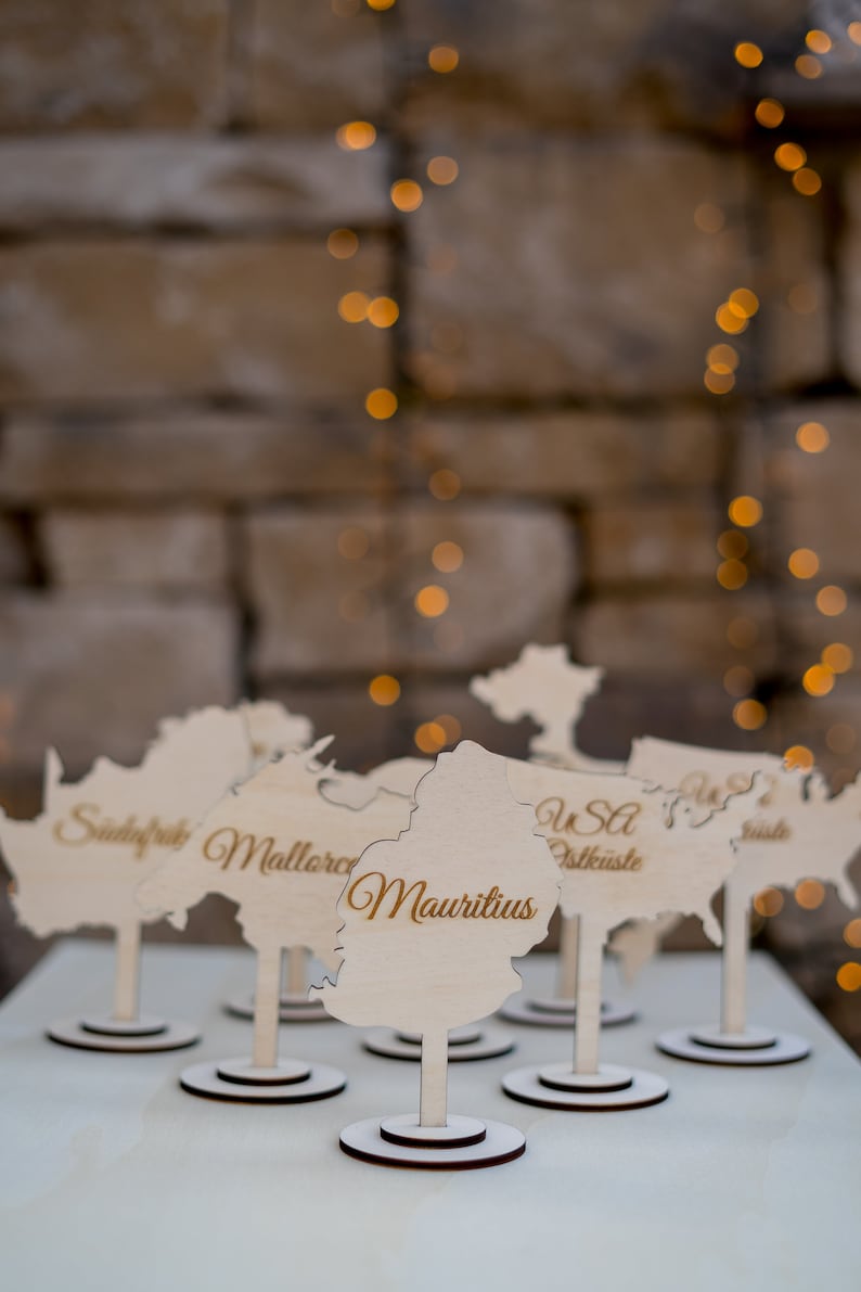 K&ouml;nnte beinhalten: Holz-Namensschilder in Form von L&auml;nderumrissen, mit Namen wie "Mauritius" und "USA Ostk&uuml;ste" in eleganter Schrift. Jede Karte steht auf einem kleinen, runden Sockel. Der Hintergrund ist eine verschwommene Backsteinmauer mit weichen, goldenen Lichtern.