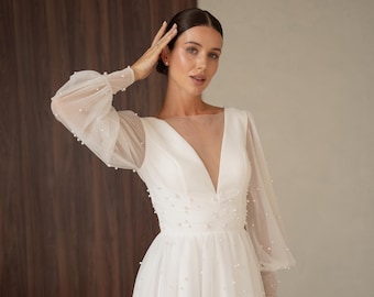 Op de afbeelding: Witte trouwjurk met een diepe V-hals en lange, doorschijnende mouwen versierd met kleine parels. De jurk heeft een getailleerd lijfje en een zwierige rok, wat een elegant silhouet creëert. De jurk wordt in een studio gedragen.