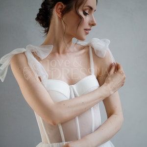 May include: A white bridal gown with a sheer bodice and a tulle skirt. The bodice has a corset-style design with boning and a sweetheart neckline. The straps are made of tulle and have large bows at the shoulders.