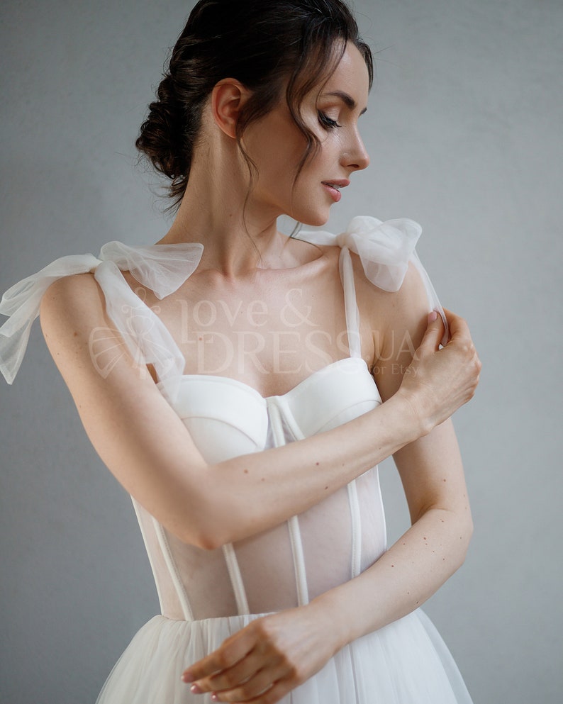 May include: A white bridal gown with a sheer bodice and a tulle skirt. The bodice has a corset-style design with boning and a sweetheart neckline. The straps are made of tulle and have large bows at the shoulders.
