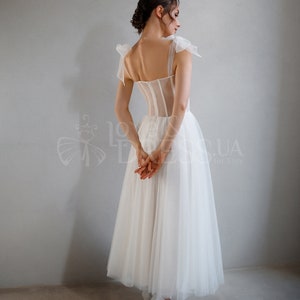 May include: A white tulle wedding dress with a sheer corset bodice and bow straps. The dress has a full skirt and is shown from the back.