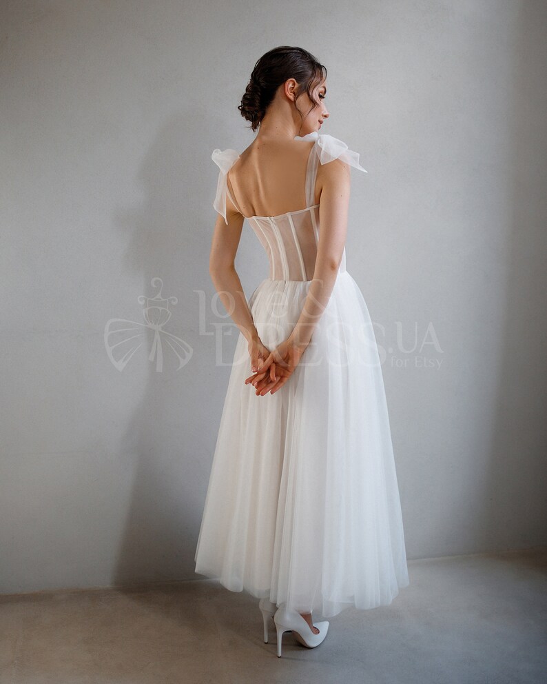 May include: A white tulle wedding dress with a sheer corset bodice and bow straps. The dress has a full skirt and is shown from the back.