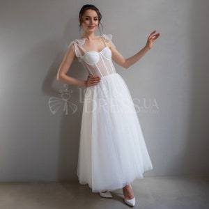 May include: A white tulle wedding dress with a sheer bodice and a sweetheart neckline. The bodice has a corset-style design with boning and a mesh overlay. The dress has a full skirt and is accented with bows on the straps.