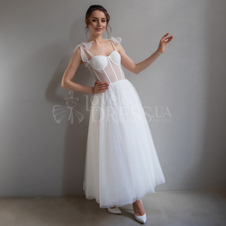 May include: A white tulle wedding dress with a sheer bodice and a sweetheart neckline. The bodice has a corset-style design with boning and a mesh overlay. The dress has a full skirt and is accented with bows on the straps.