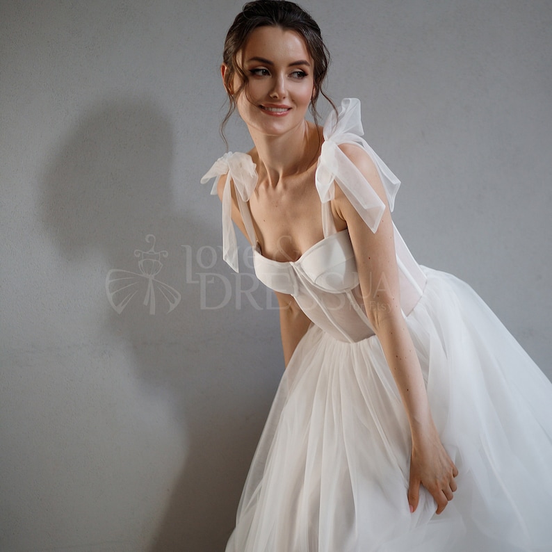 May include: A white wedding dress with a fitted bodice and a full skirt. The bodice has a sweetheart neckline and is made of satin. The skirt is made of tulle and has a sheer overlay. The dress has thin straps with bows at the shoulders.