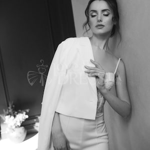 May include: A woman wearing a white pantsuit with a white blazer and a lace top. She is standing with her hand on her blazer and looking down.