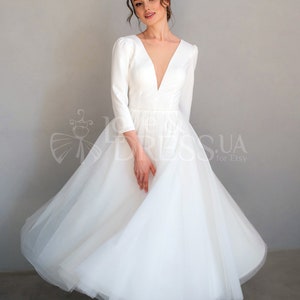 May include: A white wedding dress with a deep V-neckline and long sleeves. The skirt is made of tulle and has a full, flowing silhouette.