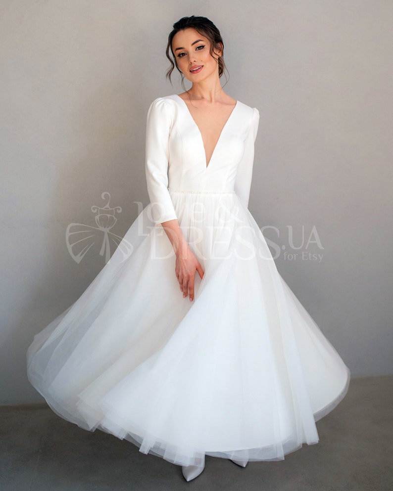 May include: A white wedding dress with a deep V-neckline and long sleeves. The skirt is made of tulle and has a full, flowing silhouette.