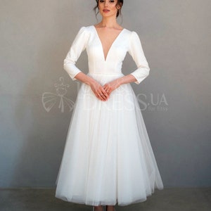 May include: A white wedding dress with a deep V-neckline, long sleeves, and a full tulle skirt. The dress has a fitted bodice and a flowing skirt.