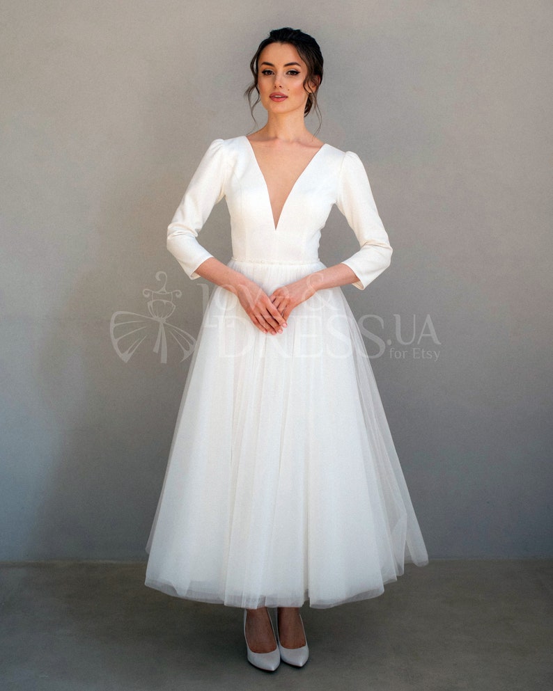 May include: A white wedding dress with a deep V-neckline, long sleeves, and a full tulle skirt. The dress has a fitted bodice and a flowing skirt.
