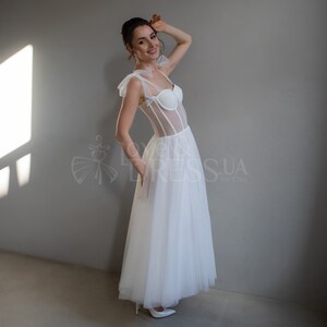 May include: A white tulle wedding dress with a sheer bodice and a corset-style top. The dress has a fitted bodice and a flowing skirt.