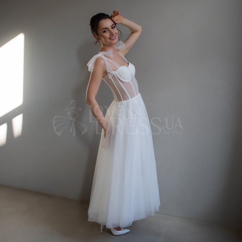 May include: A white tulle wedding dress with a sheer bodice and a corset-style top. The dress has a fitted bodice and a flowing skirt.