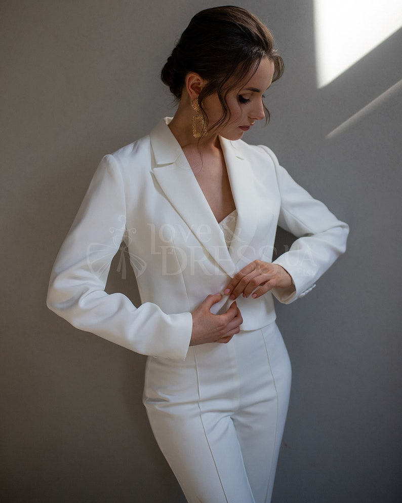 May include: A white pantsuit with a cropped blazer and a fitted silhouette. The blazer has a classic notched lapel and long sleeves. The pants have a high waist and a straight leg.