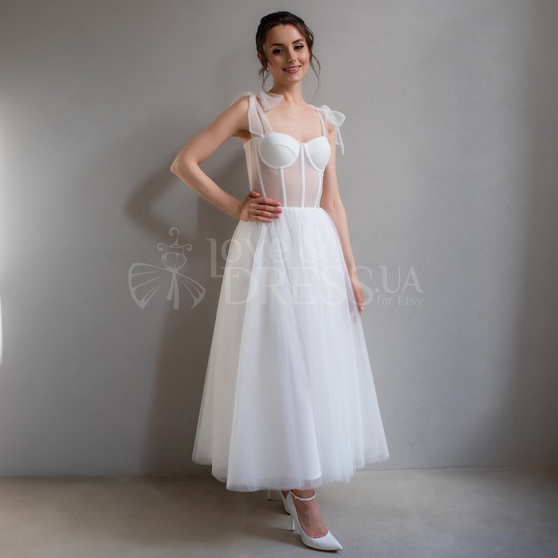 May include: A white tulle wedding dress with a fitted bodice featuring a sheer mesh panel and a sweetheart neckline. The dress has a full skirt and is accented with delicate bows on the straps.