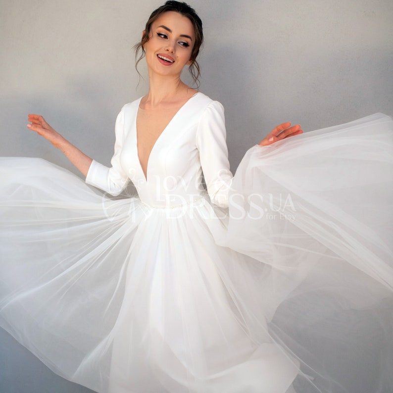 May include: A white wedding dress with a deep V-neckline and long sleeves. The dress has a tulle skirt and a fitted bodice.