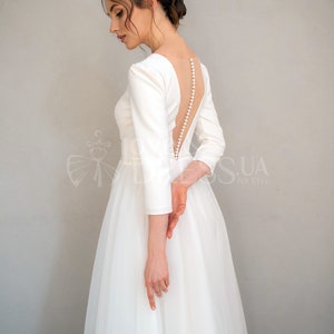 May include: A white wedding dress with a low back and a row of pearl buttons. The dress has long sleeves and a full skirt.
