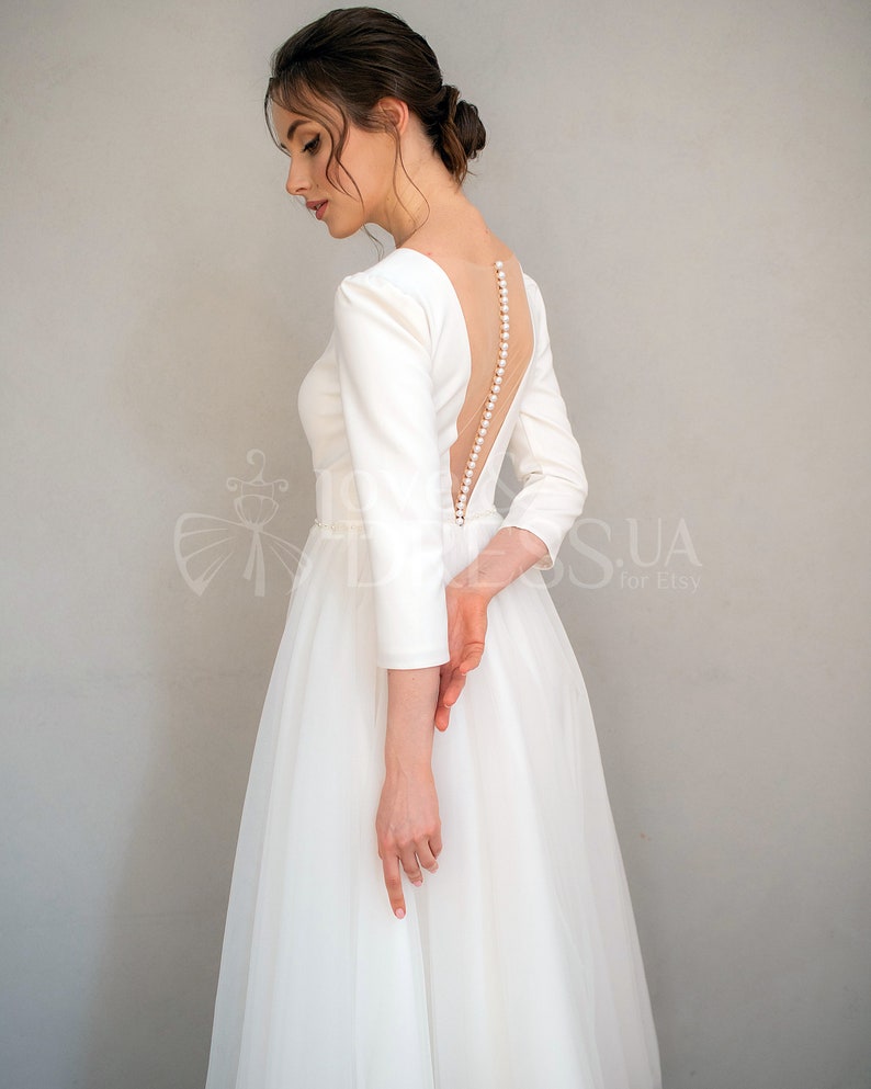 May include: A white wedding dress with a low back and a row of pearl buttons. The dress has long sleeves and a full skirt.