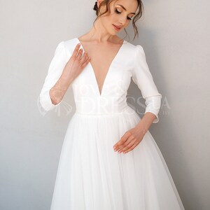 May include: A white wedding dress with a deep V-neckline and three-quarter sleeves. The dress has a fitted bodice and a flowing tulle skirt.