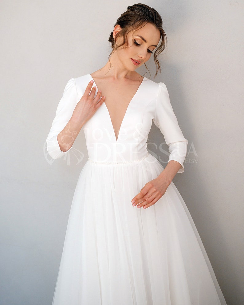 May include: A white wedding dress with a deep V-neckline and three-quarter sleeves. The dress has a fitted bodice and a flowing tulle skirt.