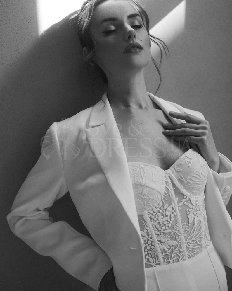 May include: A white blazer with a single button closure and a white lace corset top with floral embroidery. The model is wearing the outfit and has her eyes closed.