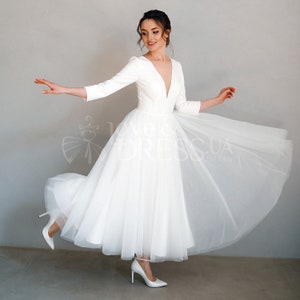 May include: A white wedding dress with a deep V-neckline, long sleeves, and a tulle skirt. The dress is fitted through the bodice and flows out into a full skirt.