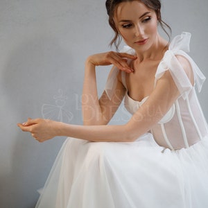 May include: A white wedding dress with a sheer bodice and a tulle skirt. The bodice has a sweetheart neckline and is adorned with delicate straps that tie into bows at the shoulders.