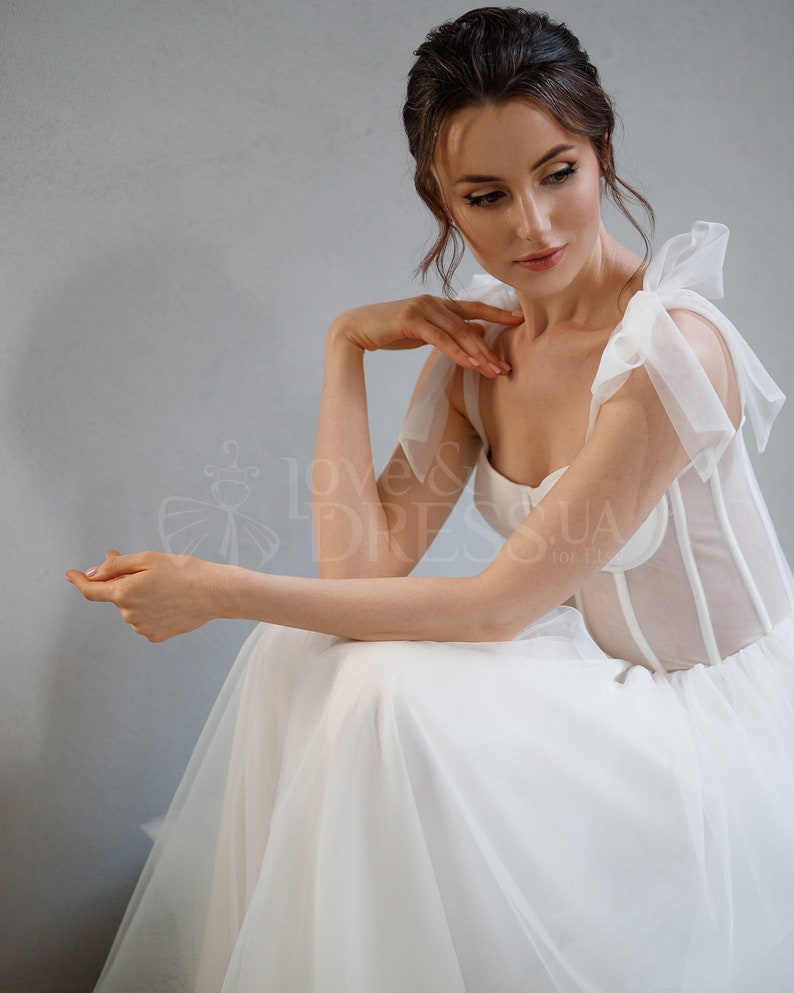 May include: A white wedding dress with a sheer bodice and a tulle skirt. The bodice has a sweetheart neckline and is adorned with delicate straps that tie into bows at the shoulders.