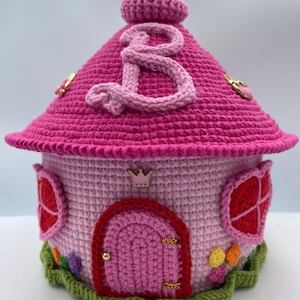 May include: A pink crocheted house with a red door, a green base, and a pink letter "B" on the roof. The house has a gold crown and other gold accents.