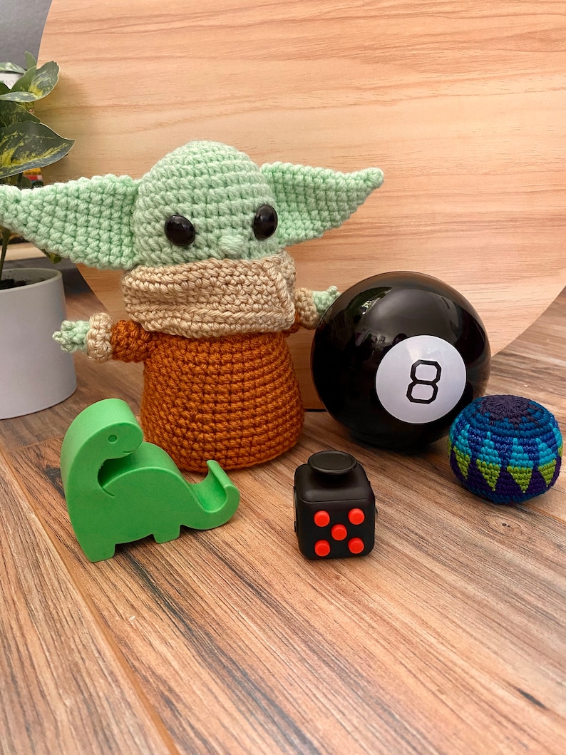 Op de afbeelding: Een groene gehaakte baby Yoda pop met een bruine gehaakte trui, een zwarte 8-bal, een groene gehaakte dinosaurus en een zwarte fidget cube met rode knoppen.