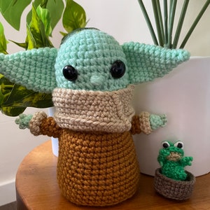 May include: A crocheted green and brown Yoda doll with large black eyes. A smaller crocheted green frog doll is sitting in a brown pot next to Yoda.
