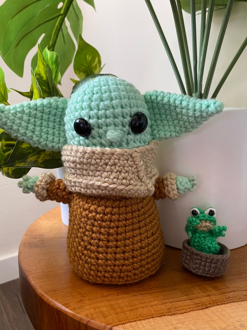 May include: A crocheted green and brown Yoda doll with large black eyes. A smaller crocheted green frog doll is sitting in a brown pot next to Yoda.
