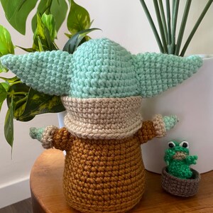 May include: A crocheted green and brown Baby Yoda doll with a small crocheted green frog doll sitting in a brown pot beside it.