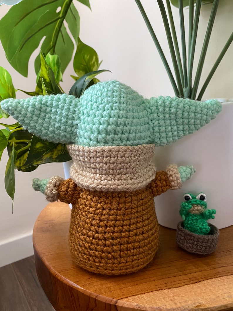 May include: A crocheted green and brown Baby Yoda doll with a small crocheted green frog doll sitting in a brown pot beside it.