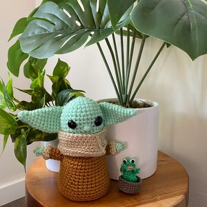 May include: A crocheted green and brown Baby Yoda doll with a tan scarf stands on a wooden table next to a small green crocheted frog in a brown pot. A large green potted plant with large leaves is in the background.
