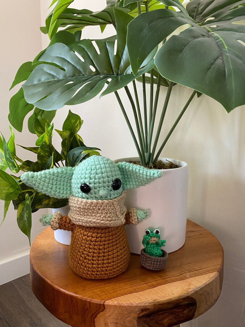May include: A crocheted green and brown Baby Yoda doll with a tan scarf stands on a wooden table next to a small green crocheted frog in a brown pot. A large green potted plant with large leaves is in the background.