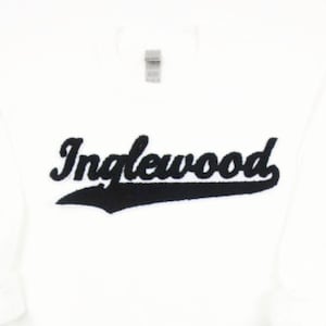 May include: White sweatshirt with the word "Inglewood" in black script across the chest. The word is slightly raised and has a black underline. The sweatshirt has long sleeves and a crew neck.